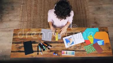 卷发妇女制作彩色拼贴画的俯视图坐在桌旁用剪刀剪人物。设计